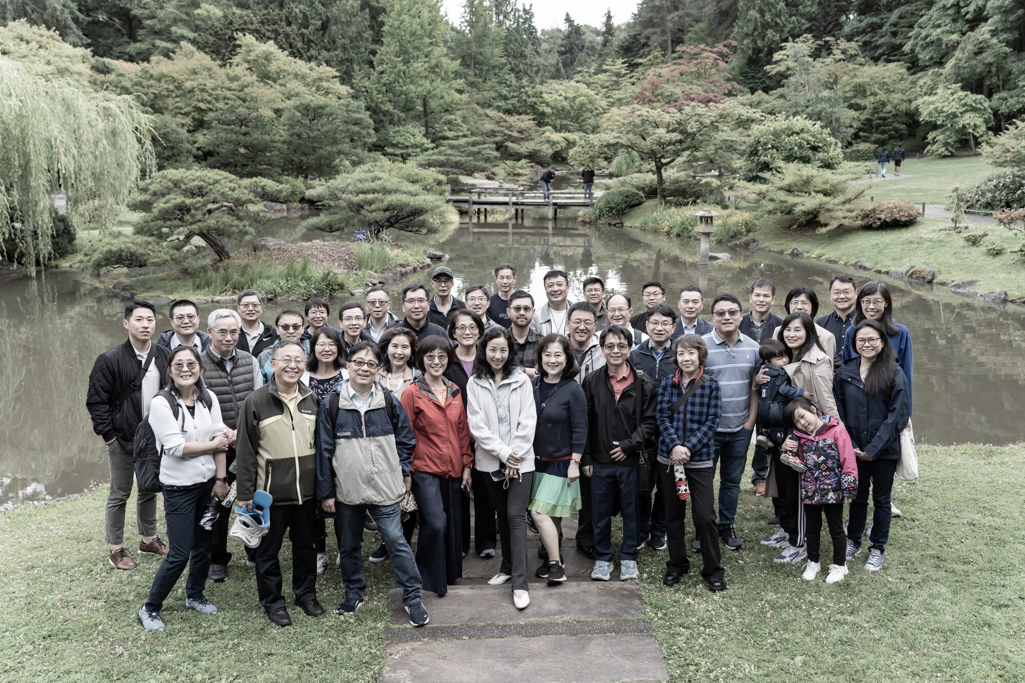 A large group stands on a lawn beside a pond in a Japanese garden.
